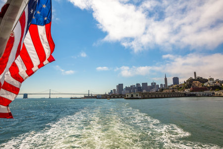 San Francisco Financial District cityscape and Oakland Bridge on sunny day, California, United States. Sea views from Alcatraz boat with American flag waving. Freedom and travel concept.の写真素材