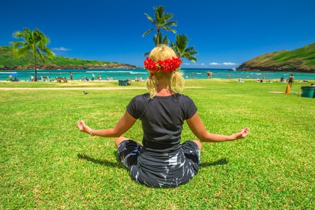 Serene meditation yoga. Woman meditating on Hawaiian palm beach in black sarong with red hawaiian lei on her head. Female contemplates ocean in popular Hanauma Bay Nature Preserve, Oahu, Hawaii, USA.の写真素材