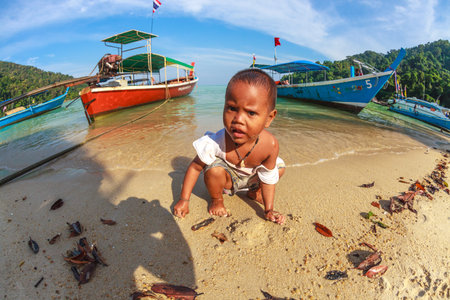 Mu Ko Surin National Park, Thailand - January 3, 2016: Sad lone child of Chao Leh or Moken tribe, playing on the beach of his village. Colored long-tail boat on background.のeditorial素材