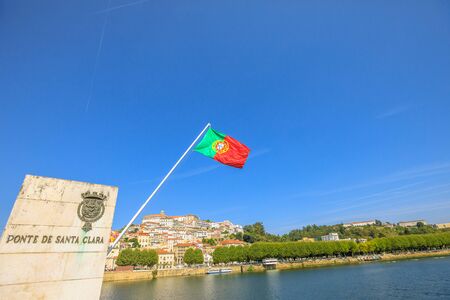 Skyline of historic city of Coimbra on Mondego river from Santa Clara bridge with flag of Portugal flying on background. Coimbra in Central Portugal, is famous for its University.の写真素材