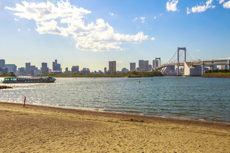 Rainbow Bridge with Tokyo Skyline from Odaiba Seaside Park in Odaiba Island, Japan. A tourist ferry connecting Tokyo with Daiba Island on background. The Rainbow Bridge is a modern and iconic landmarkの写真素材
