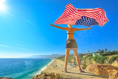 Woman holding an American flag in the blue sky from Point Dume promontory on Malibu coast in CA, United States above Point Dume Beach.Happy girl in California West Coast. Freedom and patriotic conceptの写真素材
