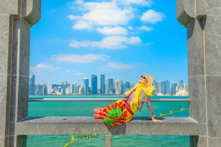 Happy blonde woman sitting under arches enjoying the view of Doha West Bay skyline outdoors of Museum of Islamic Art. Caucasian tourist in Qatar, Middle East, Arabian Peninsula. Sunny day blue sky.の写真素材