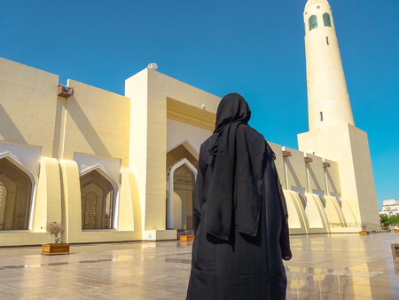 Back of woman with abaya dress looking the facade and high minaret of State Grand Mosque in Doha, Qatar, Middle East, Arabian Peninsula. Sunny day with blue sky. Muslim religion concept.の写真素材
