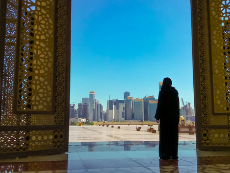 Woman with abaya dress looks at views of modern skyscrapers of Doha West Bay skyline outdoors State Grand Mosque in Doha, Qatar, Middle East, Arabian Peninsula. Sunny day with blue sky.の写真素材