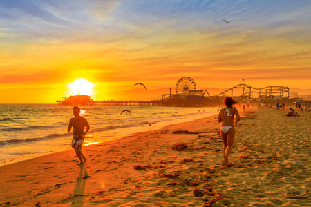 Santa Monica, California, USA - August 8, 2018: people running on seashore of Santa Monica Pier, Pacific Coast. Amusement park, ferris wheel and roller coaster. Jogging in sunset Californian beach.のeditorial素材