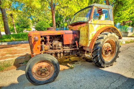 Side view of an old yellow vintage tractor with broken wheel abandoned in an old street.の写真素材