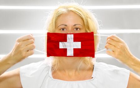 Woman on white with a Swiss flag on a surgical mask. Concept of the outbreak and quarantine for coronavirus pandemic in Switzerland. Antiviral research against the COVID-19 epidemic virus.の写真素材