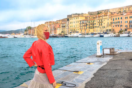 Caucasian woman with a surgical mask during Covid-19 in Portoferraio Harbour, Elba Island. Italian tourist woman travels on Elba Island. Coronavirus holiday travel in Italy with COVID pandemic.の写真素材