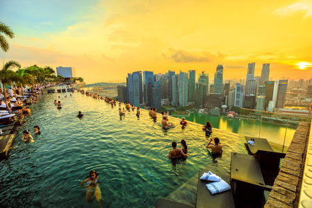 Singapore - May 3, 2018: Couples and tourists take selfie inside famous Infinity Pool of Skypark that tops the Marina Bay Sands Hotel. Financial district skyline on background. Shot at sunset.