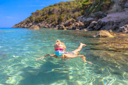 Woman swimming in Fetovaia beach and crystal sea with a surgical mask during Covid-19. Blonde tourist having a bath, Elba island, in Coronavirus quarantine. Holidays in COVID pandemic Italy.の写真素材