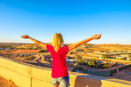 Carefree woman looking with open arms at panoramic view of Coober Pedy at sunset and surrounding desert from cave lookout. South Australian outback. Famous lookout over the city.の写真素材