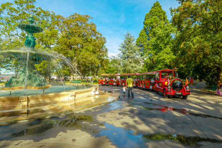 Geneva, Switzerland - Aug 15, 2020: Fountain of Four Seasons in center of Jardin Anglais with Red tourist train with carriages and locomotive along the promenade du Lac. People visit Geneva city.のeditorial素材