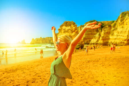 Tourism in Portugal, Europe. Lifestyle blonde female tourist on sandstone rocks of Praia Dona Ana, Lagos. Caucasian freedom woman with raised arms enjoying at Dona Ana Beach on Algarve coast.の写真素材