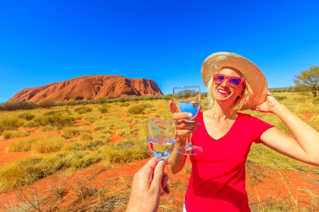 Uluru, Northern Territory, Australia - Aug 24, 2019: Lifestyle woman doing cheers to celebrate at Uluru at sunset in Uluru-Kata Tjuta National Park. Happy female in Australian outback Red Center.のeditorial素材