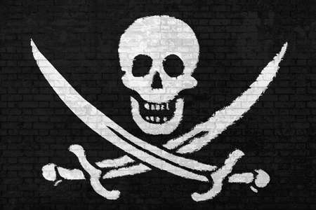 wall of bricks painted with the Pirate Flag of Calico Jack Rackham, white skull and swords crossing on black wall background. Concept of social barriers and divisions or political conflicts.の写真素材