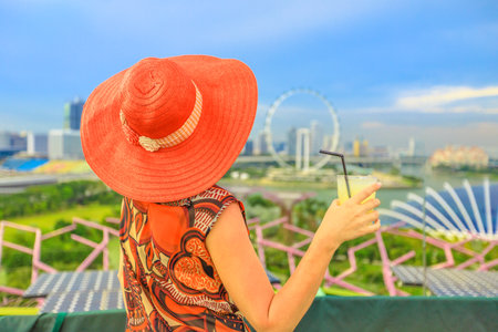 Lifestyle woman in wide hat drinking at rooftop cityscape skyline. Tourist enjoying above aerial view of Singapore marina. Travel holiday vacation in Singapore, Southeast Asia.の写真素材