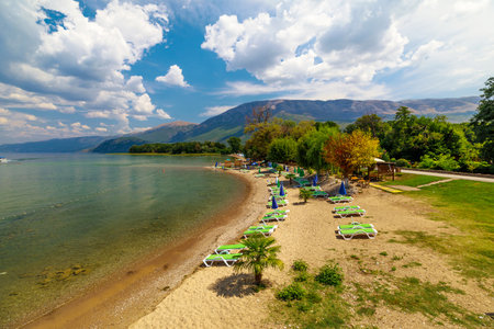 Green sunbeds and umbrellas populate the sandy beach of lake Ohrid, with crystal clear waters gently lapping the shore under a bright summer sky in North Macedoniaの写真素材