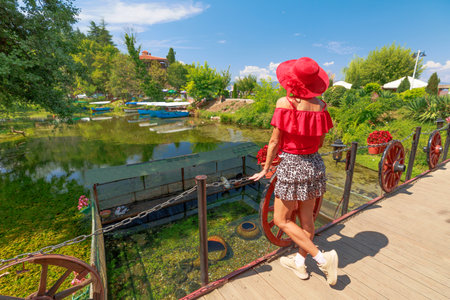 Tourist in a red shirt and wide brimmed hat walks on a wooden bridge with old wagon wheels in a lush park setting of springs in lake Ohrid, North Macedoniaの写真素材