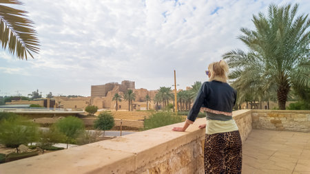 Blonde tourist admiring the landscape of the ancient mud-brick city of Diriyah, a UNESCO World Heritage site, located near Riyadh, Saudi Arabia, surrounded by palm trees and a cloudy skyの写真素材