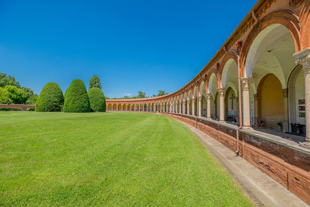 Ancient cemetery cloister featuring hanging lamps, tombs, and sculptures in the 15th-century Charterhouse of Ferrara, set against a lush green lawn, in Ferrara city of Italyの写真素材
