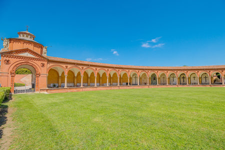 in Ferrara city of Italy, the ancient cemetery cloister featuring archway, tombs, and sculptures in the 15th-century Charterhouse of Ferrara, set against a lush green lawn with San Cristoforo churchの写真素材