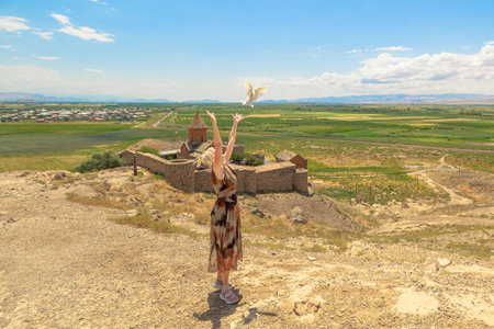 Blonde woman in long dress releasing a white dove with outstretched arms, set against the Khor Virap monastery and panoramic Ararat plain view in Armeniaの写真素材