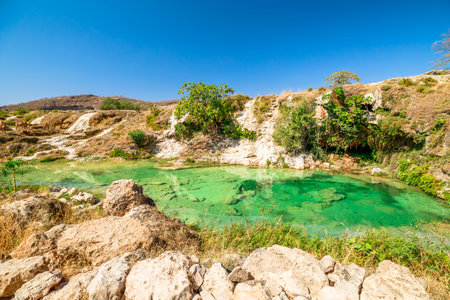 Wadi Darbat natural park in Dhofar, Oman, presenting clear emerald water with waterfalls cascading over lush rocks and green trees under a bright blue skyの写真素材