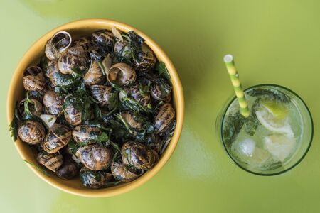 Top view of garden snails cooked French style in butter with parsley and garlic, in a yellow ceramic bowl with a glass of Mojito on a green tableの写真素材