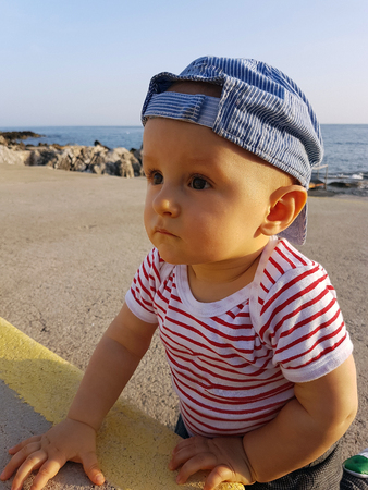 Cute Baby Boy 1 Year Wearing A Cap Backwards And Striped Clothes, Close Up Portrait. Mediterranean Sea In The Backgroundの写真素材
