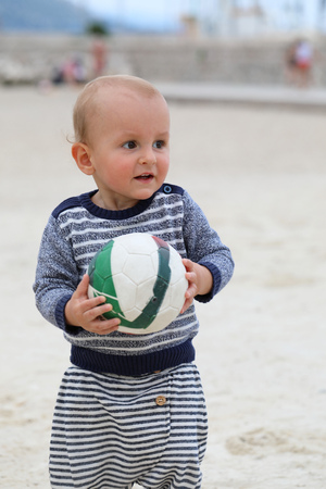 Adorable Blond Baby Boy is Holding a Soccer Ball on Sand on Sea Beachの写真素材
