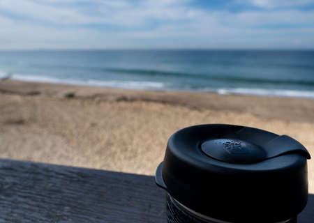 NEWCASTLE, AUSTRALIA - June 16 2019 Close up of a glass Keepcup with black trimming, filled with coffee, on a timber bench with the Pacific Ocean in the backgroundのeditorial素材