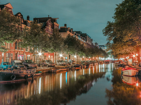 Small boats docked along a canal in Amsterdam at night, illuminated by street lights with buildings reflecting off the still water.のeditorial素材