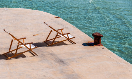 Beach deck chairs facing the sun at turquoise sea and rusty bollard at exclusive spot