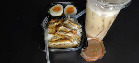 Fried pork with rice and soft boiled egg in a black box Comes with spicy dipping sauce and iced coffee on a black wooden floor. from top viewの写真素材