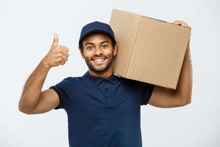 Delivery Concept - Portrait of Happy African American delivery man holding a box package and showing thumps up. Isolated on Grey studio Background. Copy Space.の写真素材