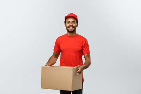 Delivery Concept - Portrait of Happy African American delivery man in red cloth holding a box package. Isolated on Grey studio Background. Copy Space.の写真素材