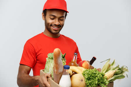 Delivery Concept - Handsome African American delivery man carrying package box of grocery food and drink from store. Isolated on Grey studio Background. Copy Space.の写真素材