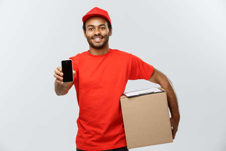 Delivery Concept - Portrait of Handsome African American delivery man or courier with box showing mobile phone on you to check the order. Isolated on Grey studio Background. Copy Space.の写真素材