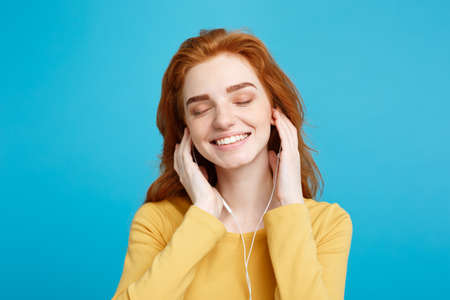 Lifestyle concept - Portrait of cheerful happy ginger red hair girl enjoy listening to music with headphones joyful smiling to camera. Isolated on Blue Pastel Background. Copy space.の写真素材