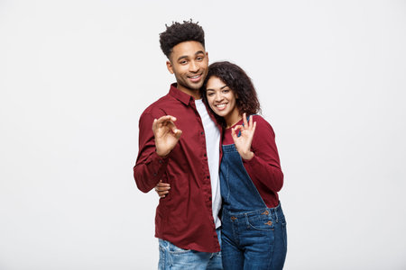 Portrait of a cheerful young african couple standing together and showing ok gesture isolated over...