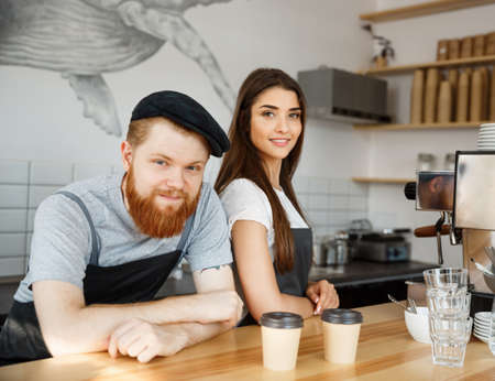 Coffee Business Concept - Positive young bearded man and beautiful attractive lady barista couple in apron looking at camera while standing at bar Counterの写真素材