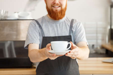 Coffee Business owner Concept - Portrait of happy young bearded caucasian barista in apron with confident looking servicing hot coffee to customer in coffee shop counter.の写真素材