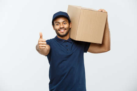 Delivery Concept - Portrait of Happy African American delivery man holding a box package and showing thumps up. Isolated on Grey studio Background. Copy Space.の写真素材