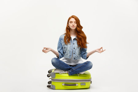 Traveling concept. Young pretty ginger woman meditating in lotus pose on the luggage valise. Isolated on white.の写真素材