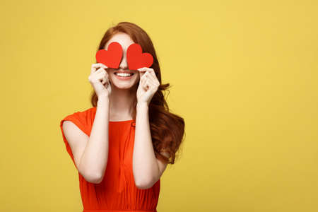 Lifestyle Concept: Attractive woman with beaming smile having two small red hearts in hands, closing eyes with paper heart symbols while standing over yellow background.の写真素材