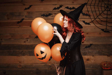 Halloween Witch Concept - Beautiful caucasian woman in witch costumes celebrating Halloween posing with posing with orange and black balloon over bats and spider web on Wooden studio background.の写真素材