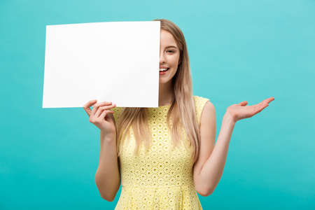 Lifestyle Concept: young beautiful girl smiling and holding a blank sheet of paper, dressed in yellow, isolated on pastel blue backgroundの写真素材