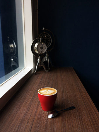Red cup of cappuccino coffee in the morning time with vintage clock background.