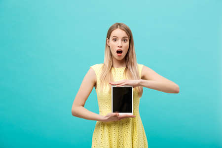 Excited shocked good-looking young woman in yellow dress having confused or puzzled expression on face while receiving urgent message or email, using wi-fi on tablet.の写真素材